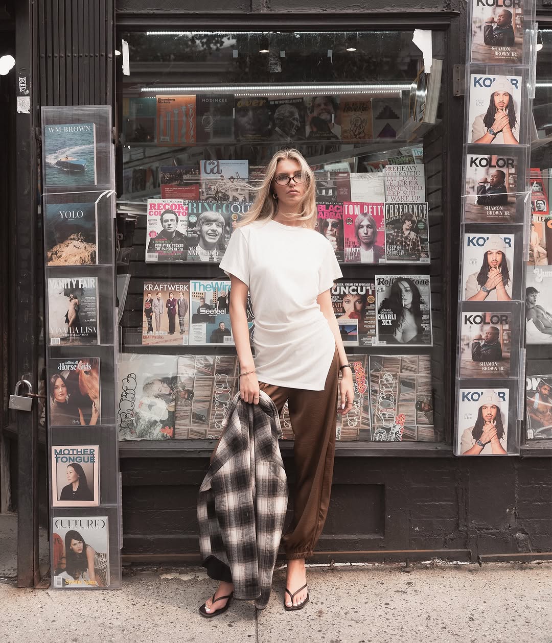 Full-body shot in front of a magazine stand featuring the same white tunic, brown pants, and held plaid shirt.