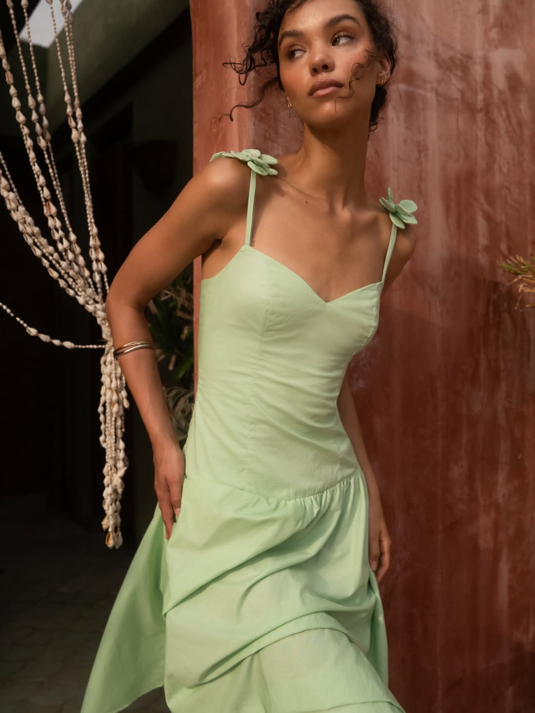 Model wearing a light green midi dress with spaghetti straps adorned with fabric flowers and a gathered skirt.