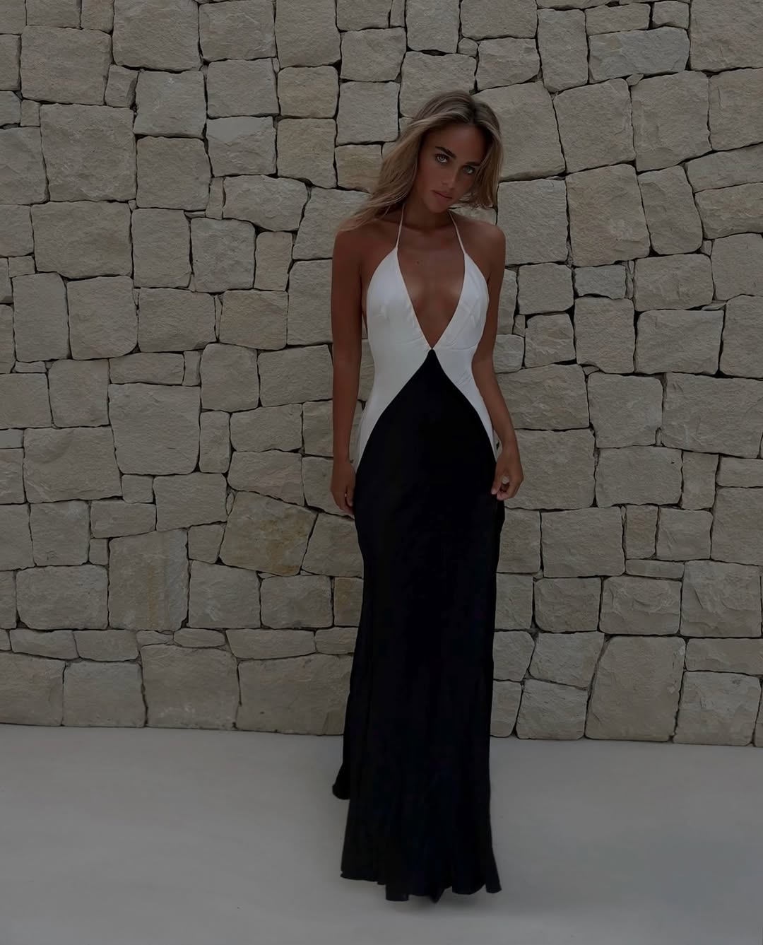 Full-body shot of the model standing in the same black and white halter gown against a stone wall.