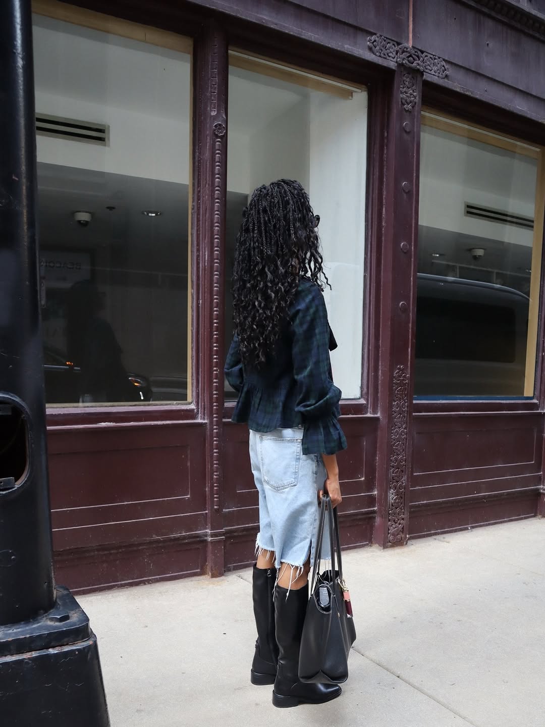 Rear view showing the back of the plaid top and denim shorts while holding a large tote bag.