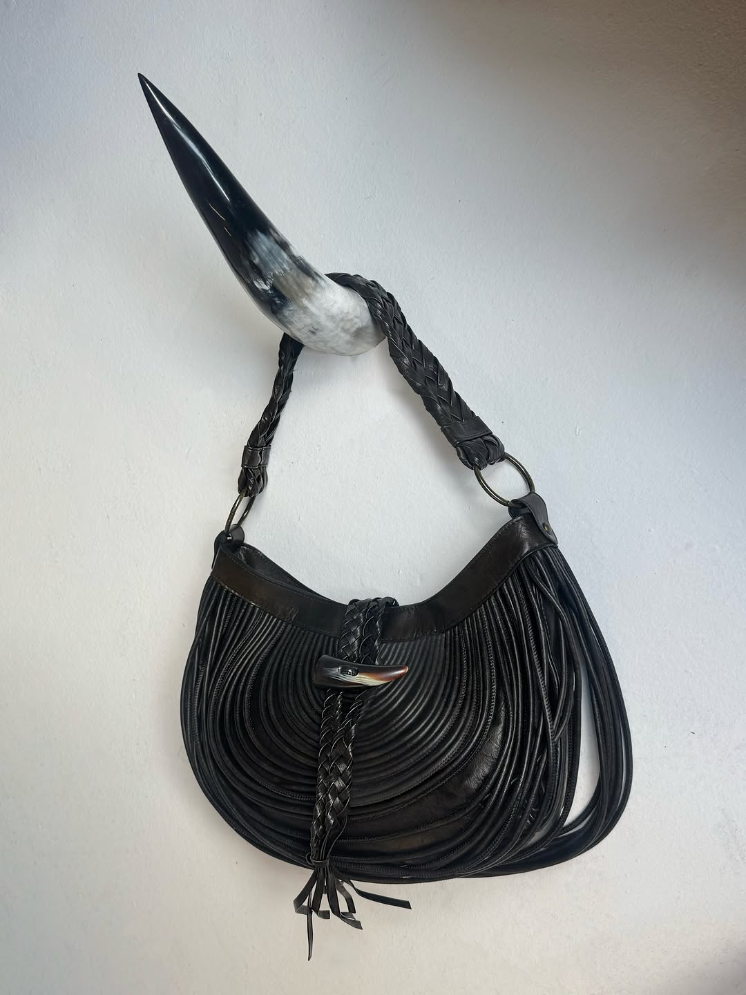 Product shot of the black fringed bag hanging on a horn-shaped wall hook against a white background.