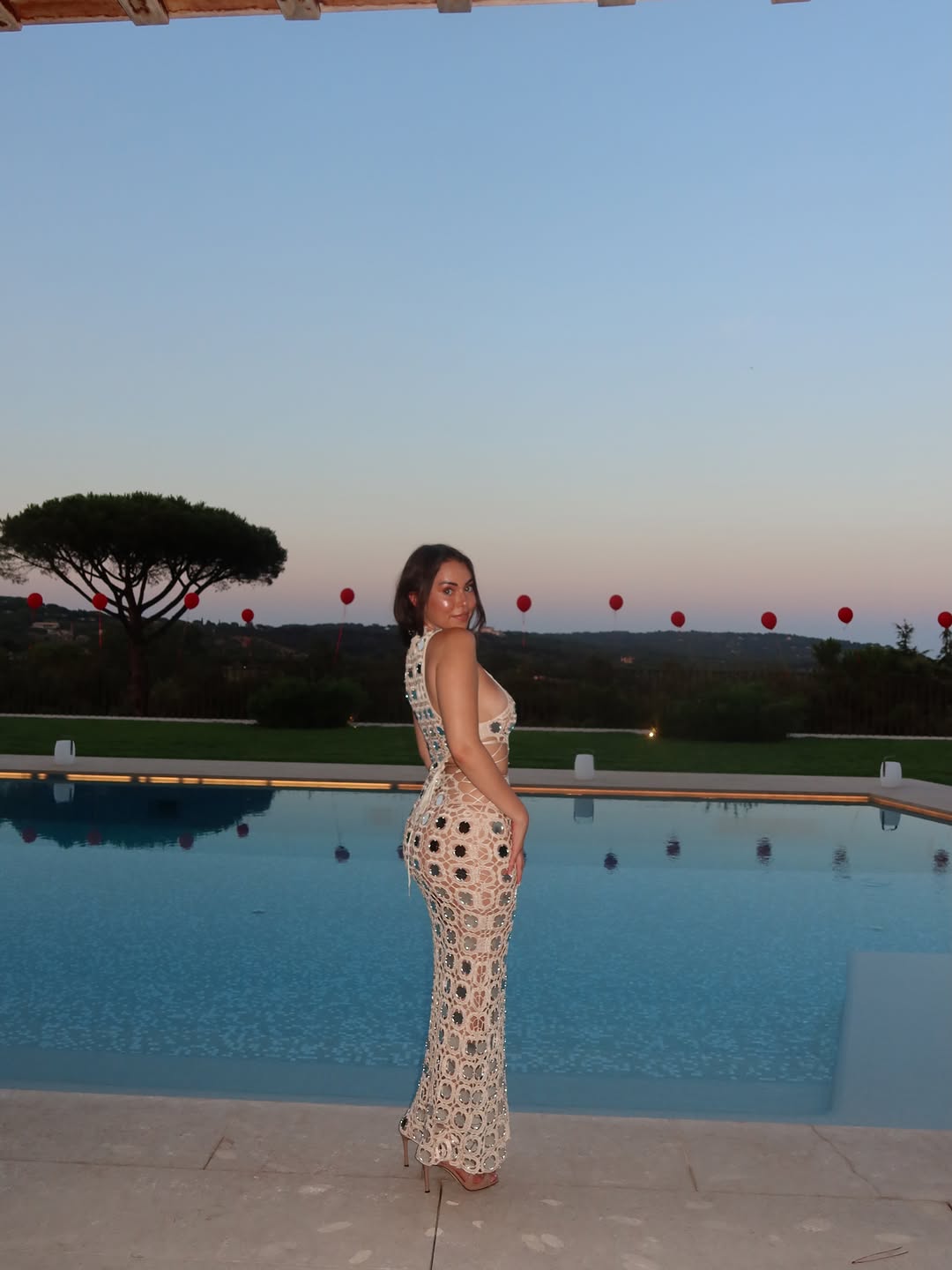 Side profile of the model standing by a pool at dusk, showing the backless design of the dress.