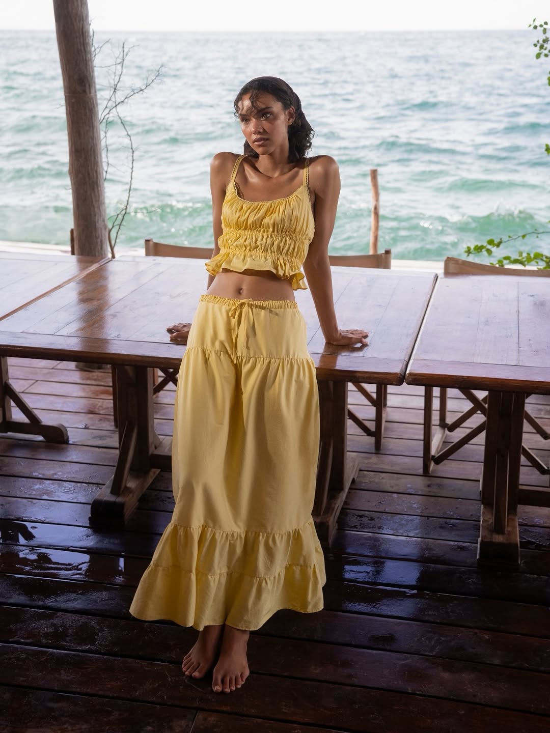 Full-body shot of the same model in the yellow two-piece set, showing the long tiered maxi skirt with ruffles at the bottom worn by the ocean.
