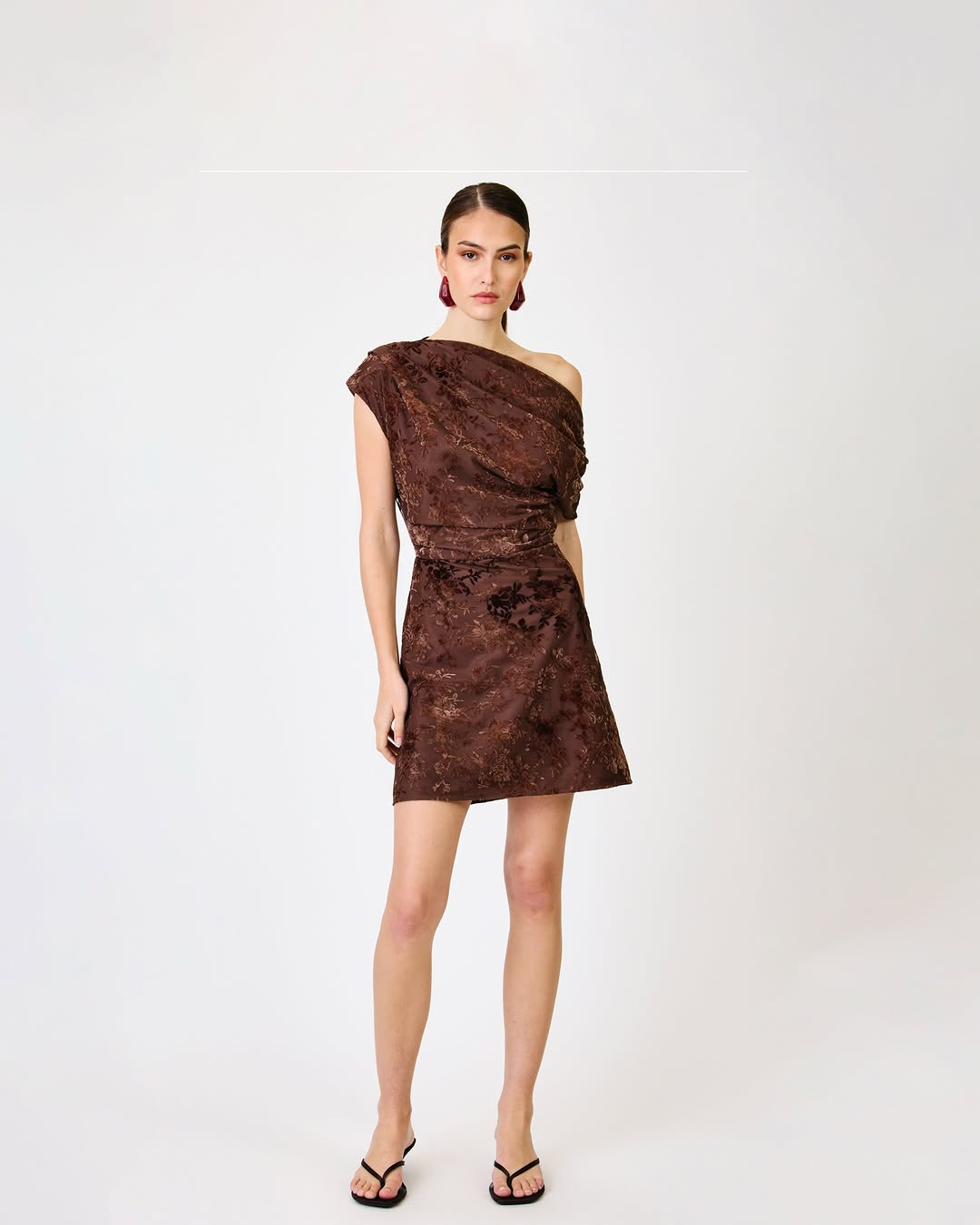 Full-body shot of the same chocolate brown floral one-shoulder mini dress styled with black thong sandals.