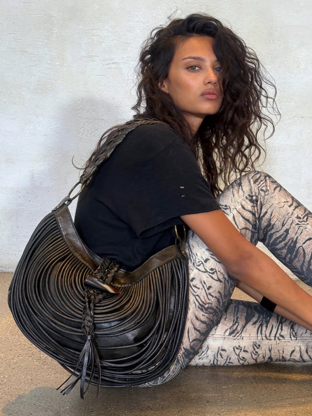 Side profile view of the model sitting down, showcasing the full silhouette of the fringed bag.