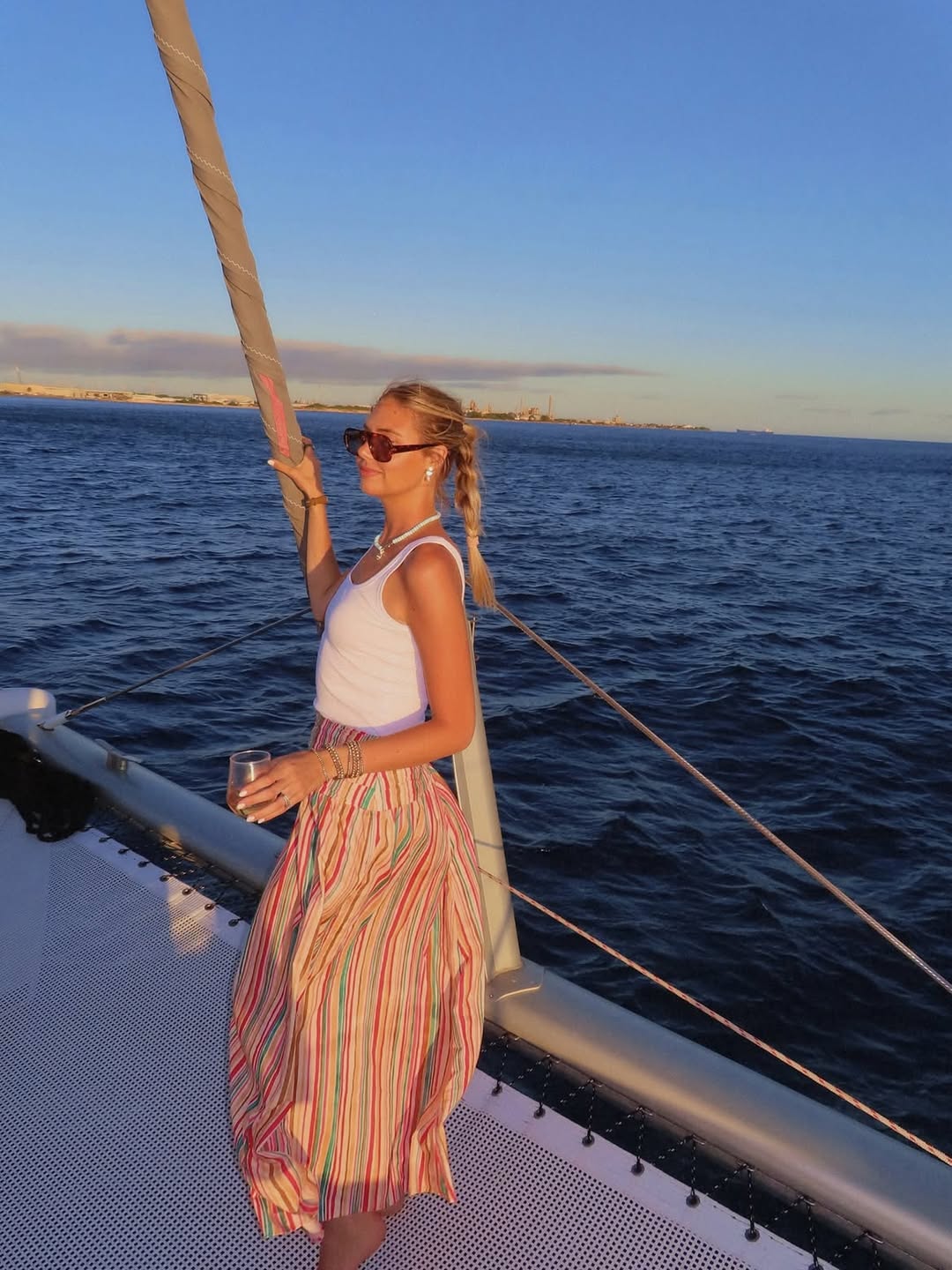 Full-body shot on a boat showing the same white tank top and striped midi skirt outfit.
