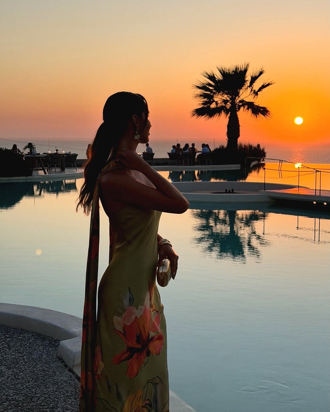 Back/side silhouette of the model in the olive green floral dress near a pool at sunset.
