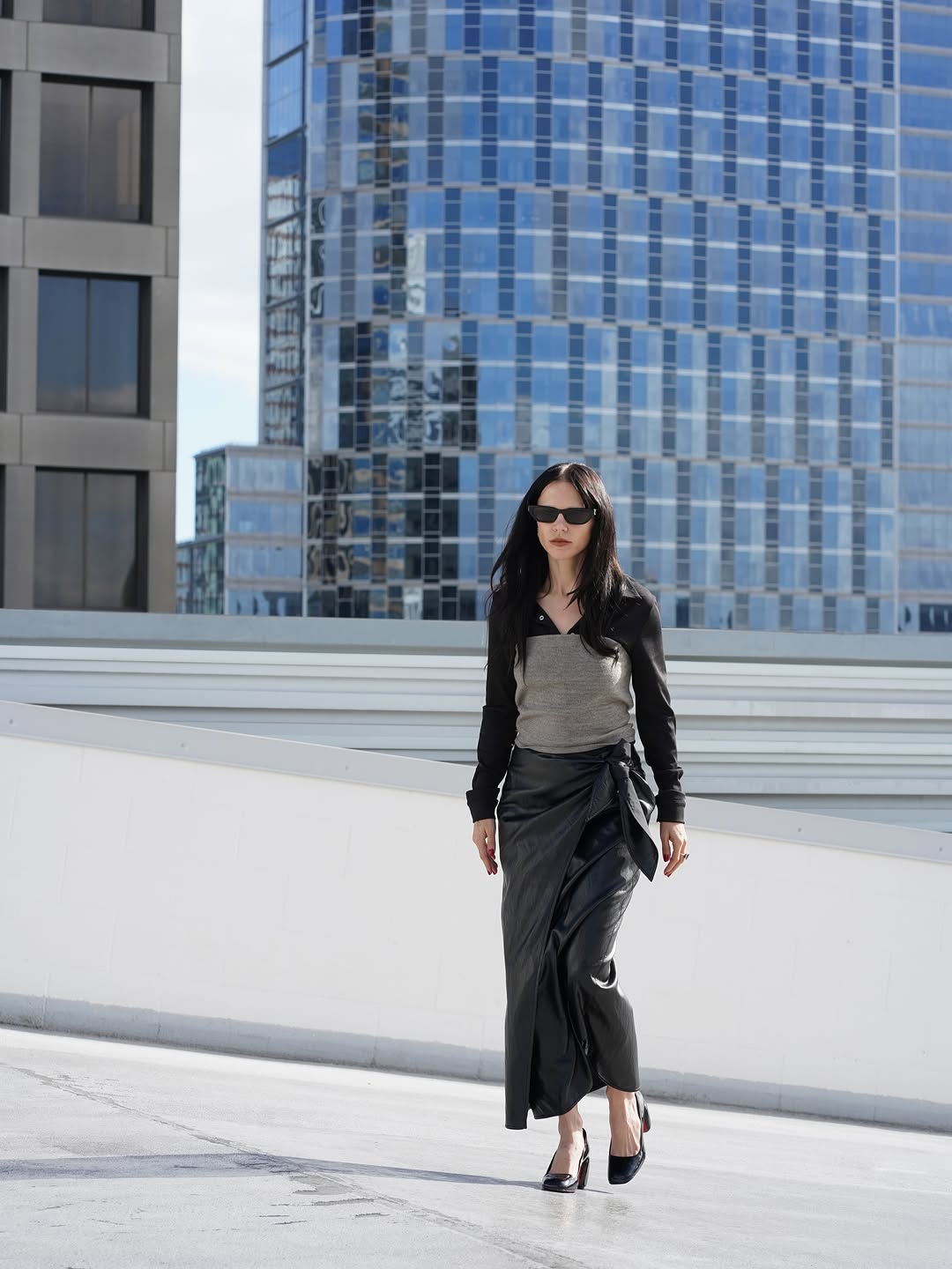 Full-body shot of the same outfit: black sheer shirt over grey tank top and black faux leather wrap skirt, styled with sunglasses and heels.