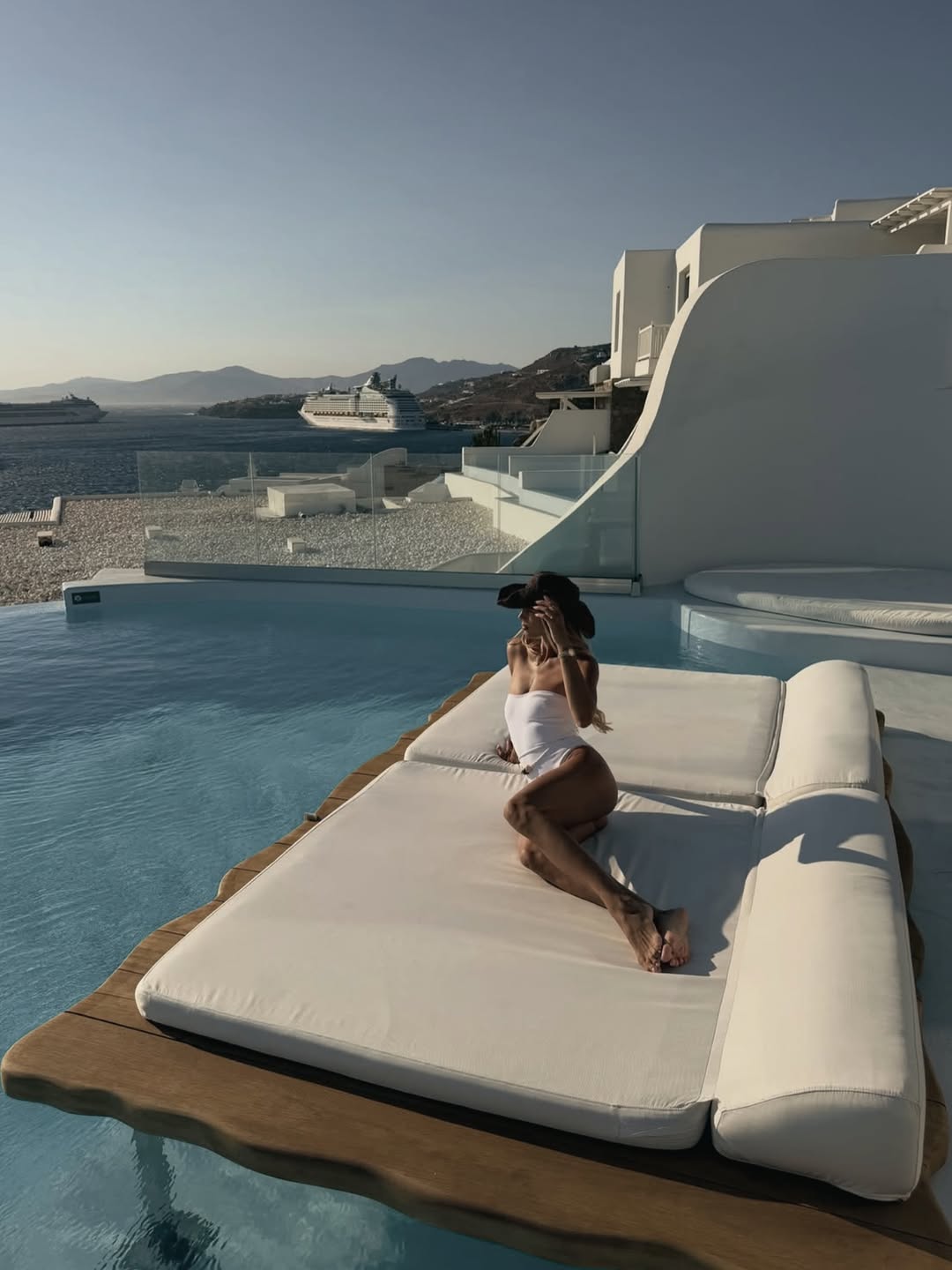 Wide shot of the model sitting on a floating daybed by an infinity pool, wearing the same white swimsuit and hat.