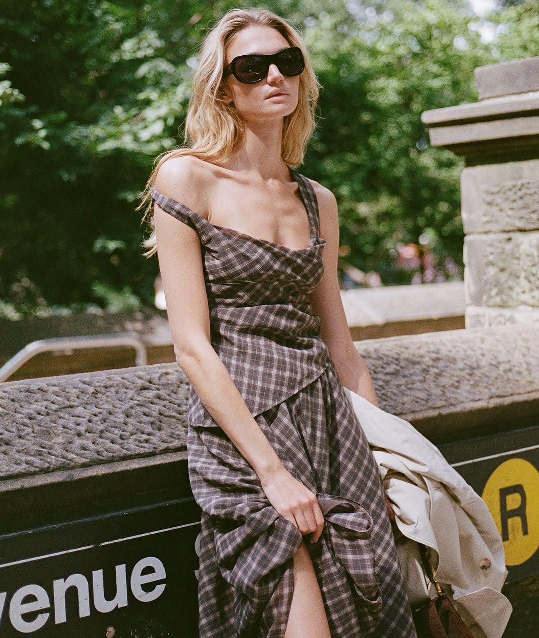 Medium shot of the same woman leaning against a stone wall, wearing the same brown plaid two-piece set with one strap pulled off her shoulder.