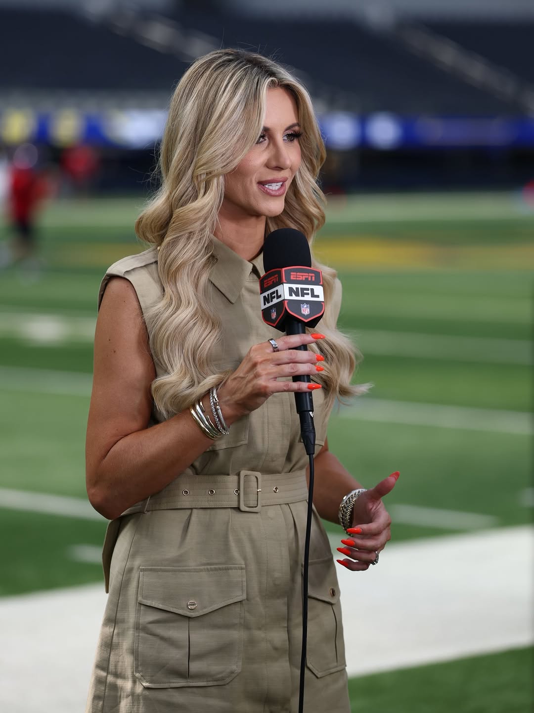 The same woman stands on a football field holding a microphone, wearing the same sleeveless beige belted dress with visible cargo pockets.