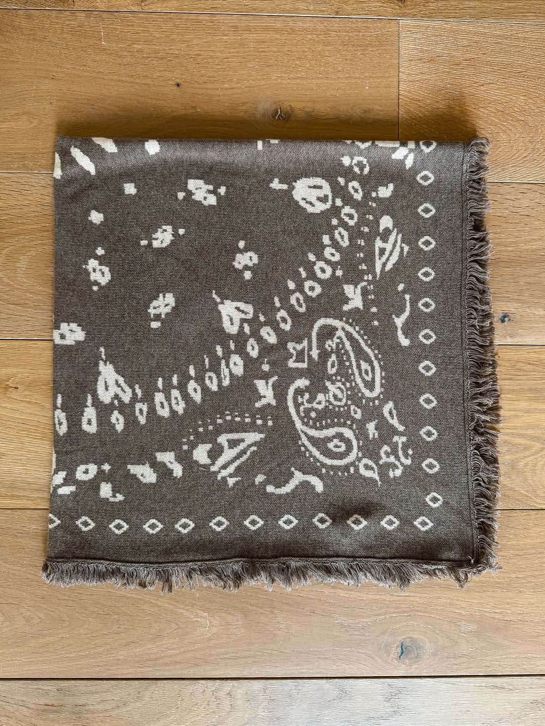 Flat lay photograph of the square brown scarf featuring white paisley patterns and fringed edges on a wooden floor.