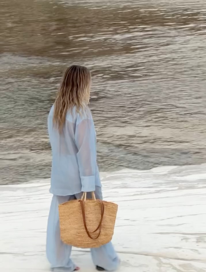 Rear view of the model in the same light blue sheer shirt and trousers set, carrying a woven tote bag.