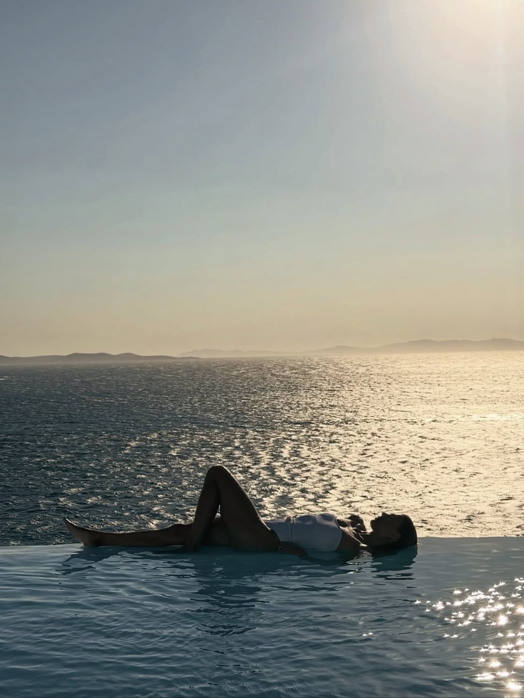 Silhouette shot of the model lying back in the water at sunset, wearing the white swimsuit.