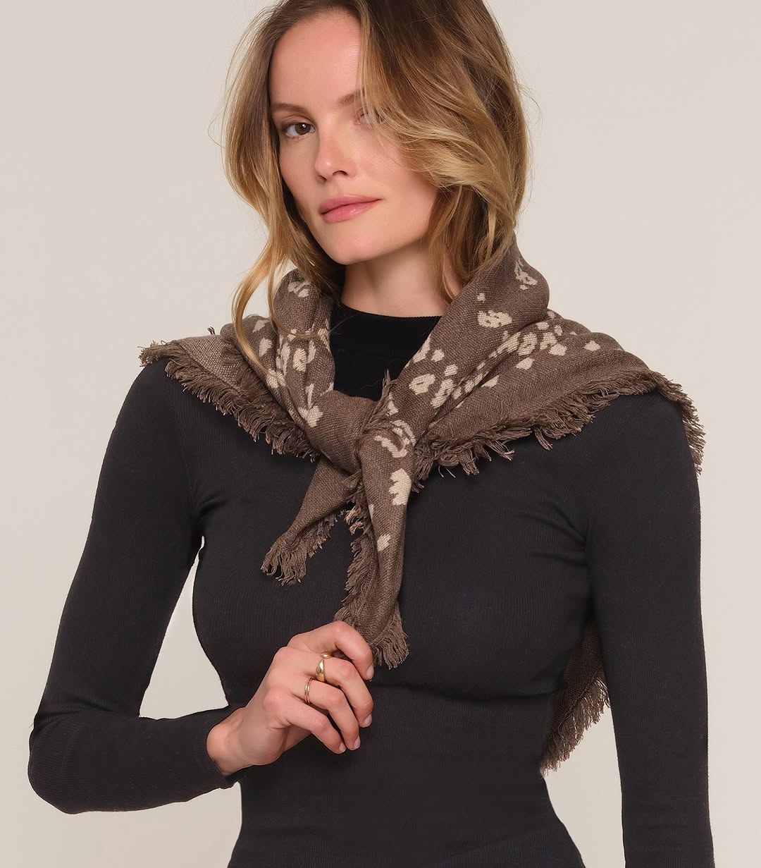 A model wearing the brown patterned fringed scarf tied around her shoulders like a shawl over a black long-sleeve top.