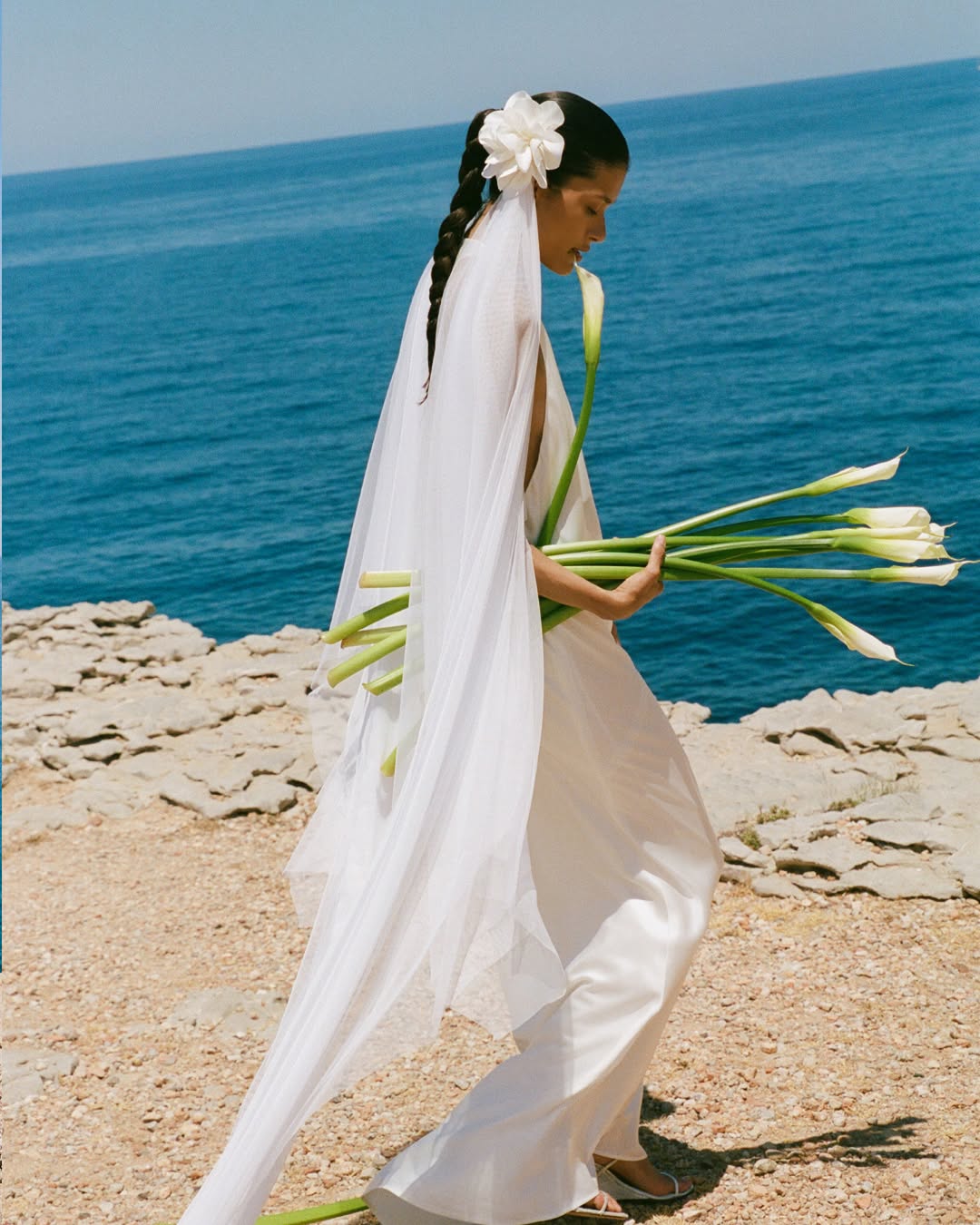 Side profile of the model in the same white gown, now styled with a long sheer veil and flower hairpiece.