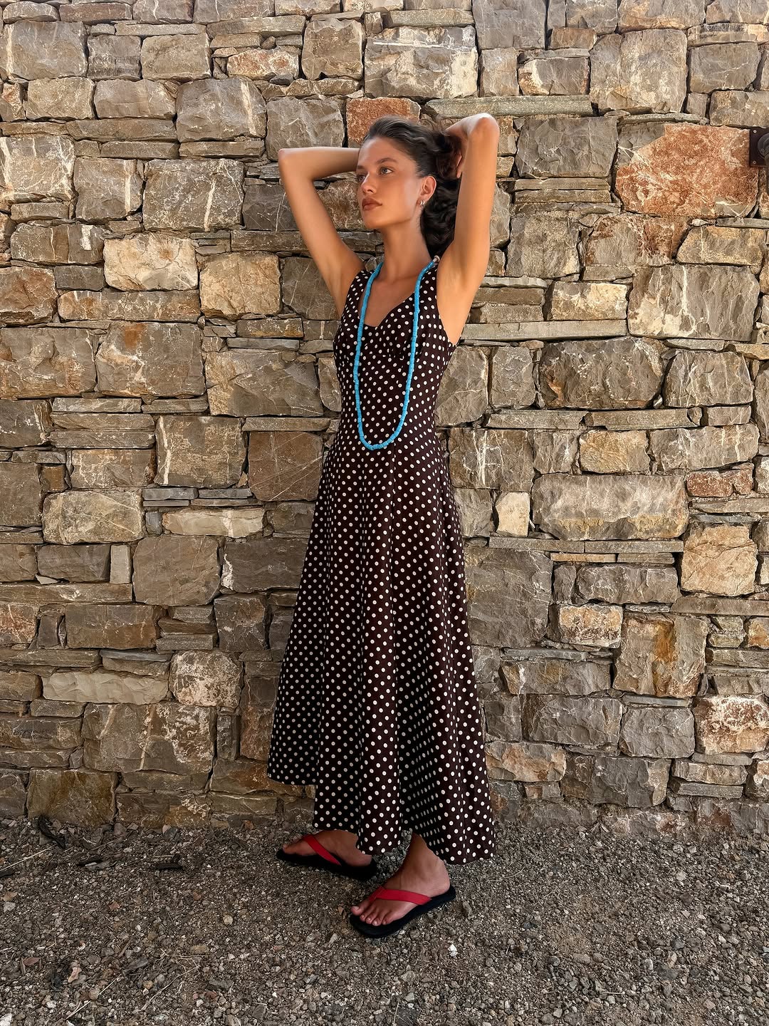 The same brown polka dot dress is shown on a standing model against a stone wall.