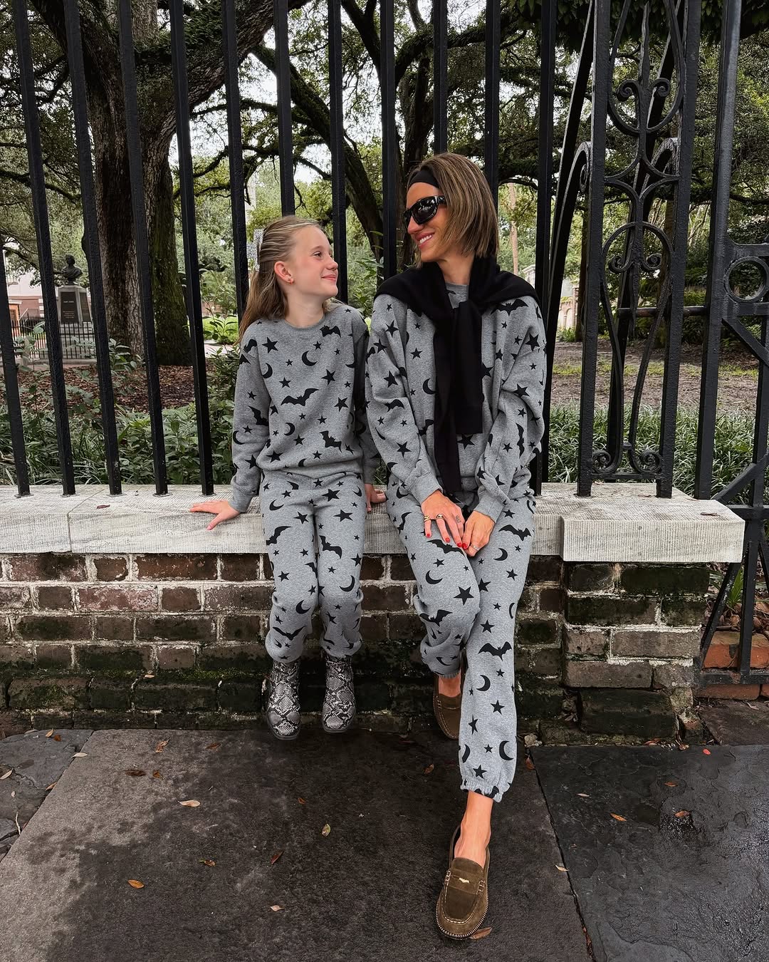 The pair sits on a low brick wall, showcasing the full fit of the matching graphic sweatshirt and jogger sets.