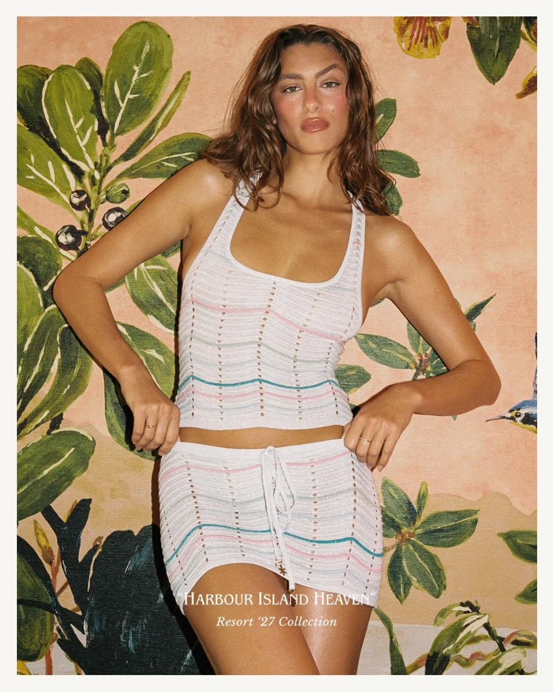 Full-body shot of the model standing against a floral backdrop wearing the white striped knit tank and mini skirt set.