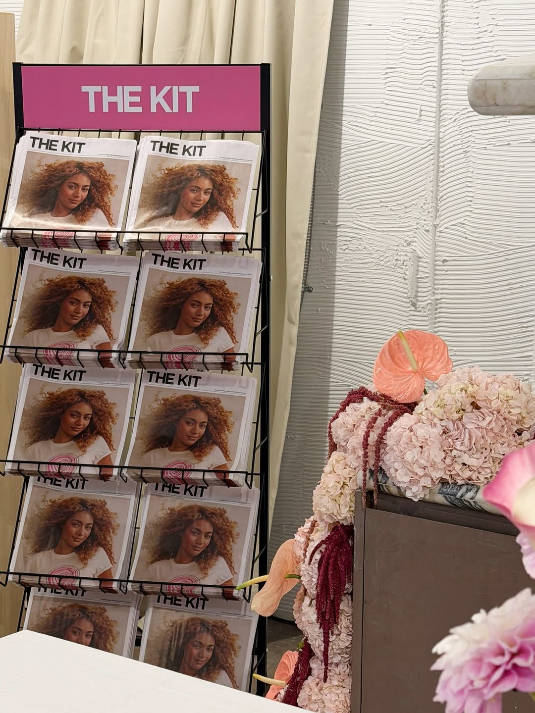 A rack displaying copies of 'THE KIT' magazine next to floral arrangements.