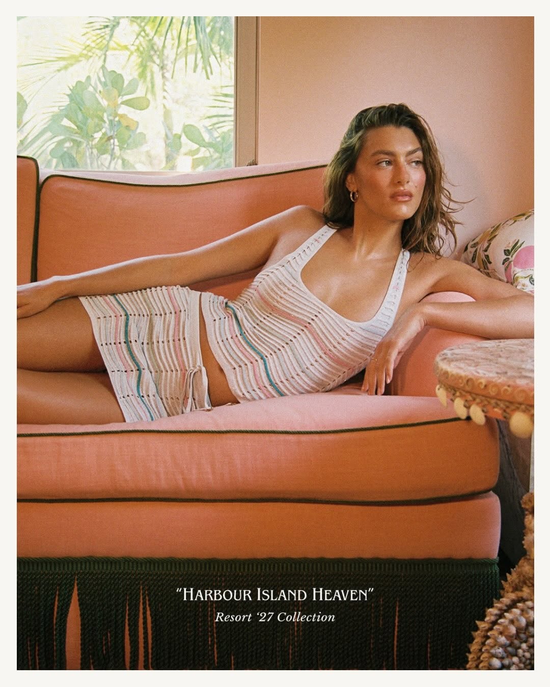 Model reclining on a pink sofa wearing the same white striped knit tank and mini skirt set.