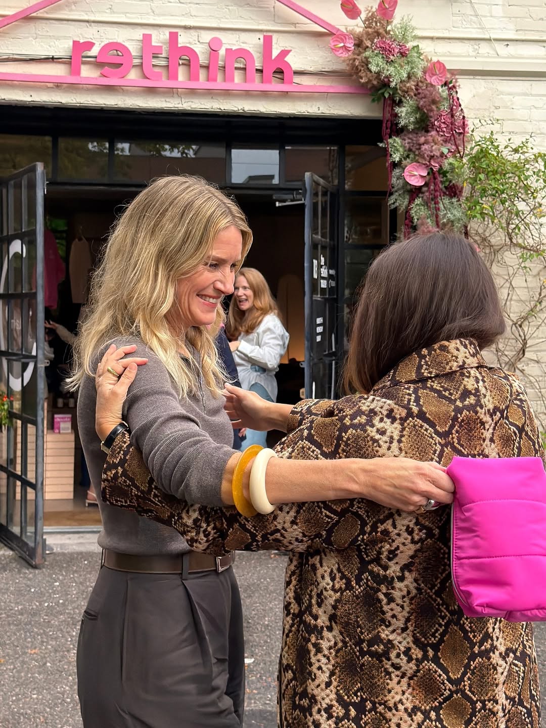 Two women hugging outside the shop; one is carrying the pink pouch under her arm.