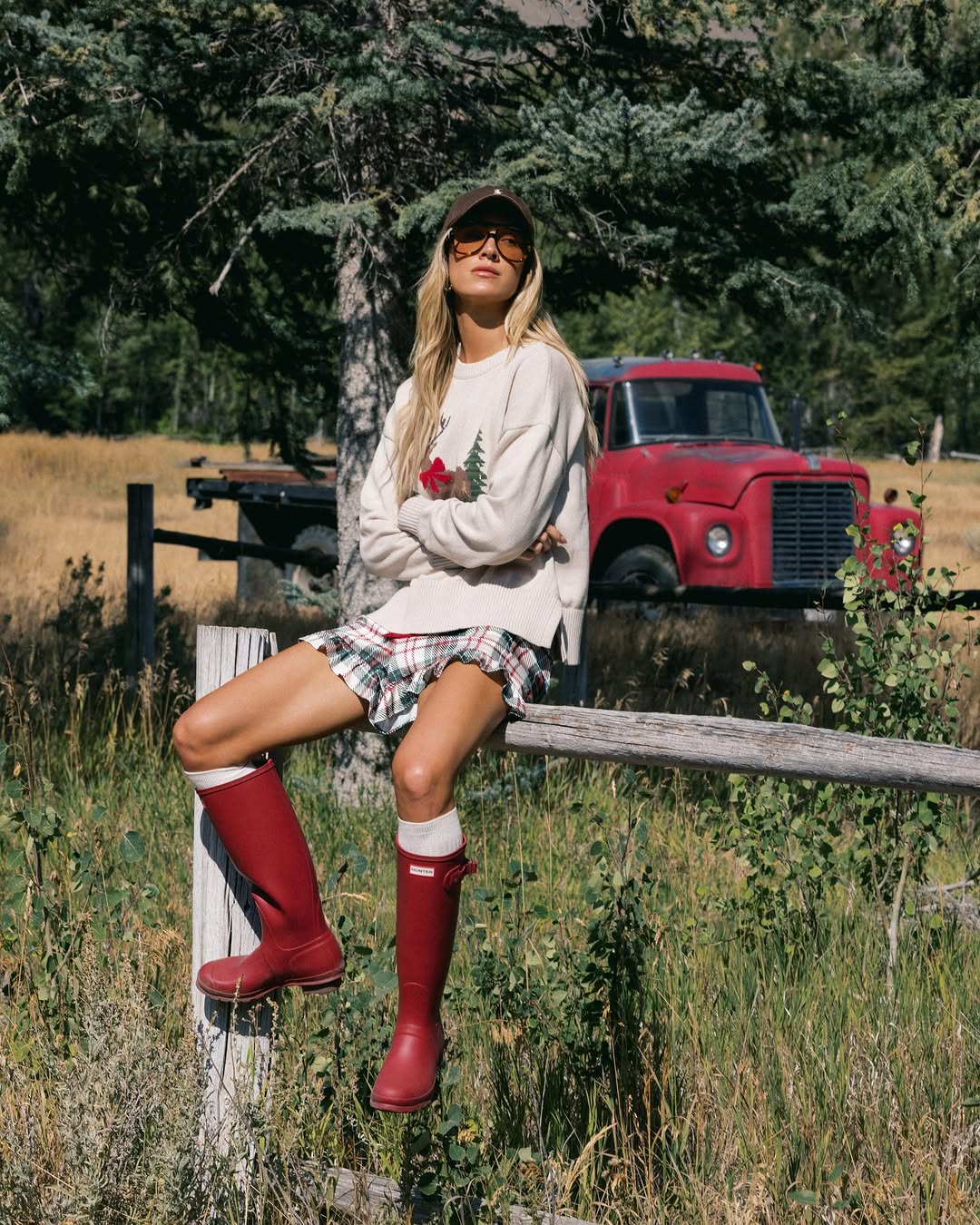 Full body shot of the model sitting on a fence post, showcasing the same sweater, plaid skirt, and tall red rubber boots.